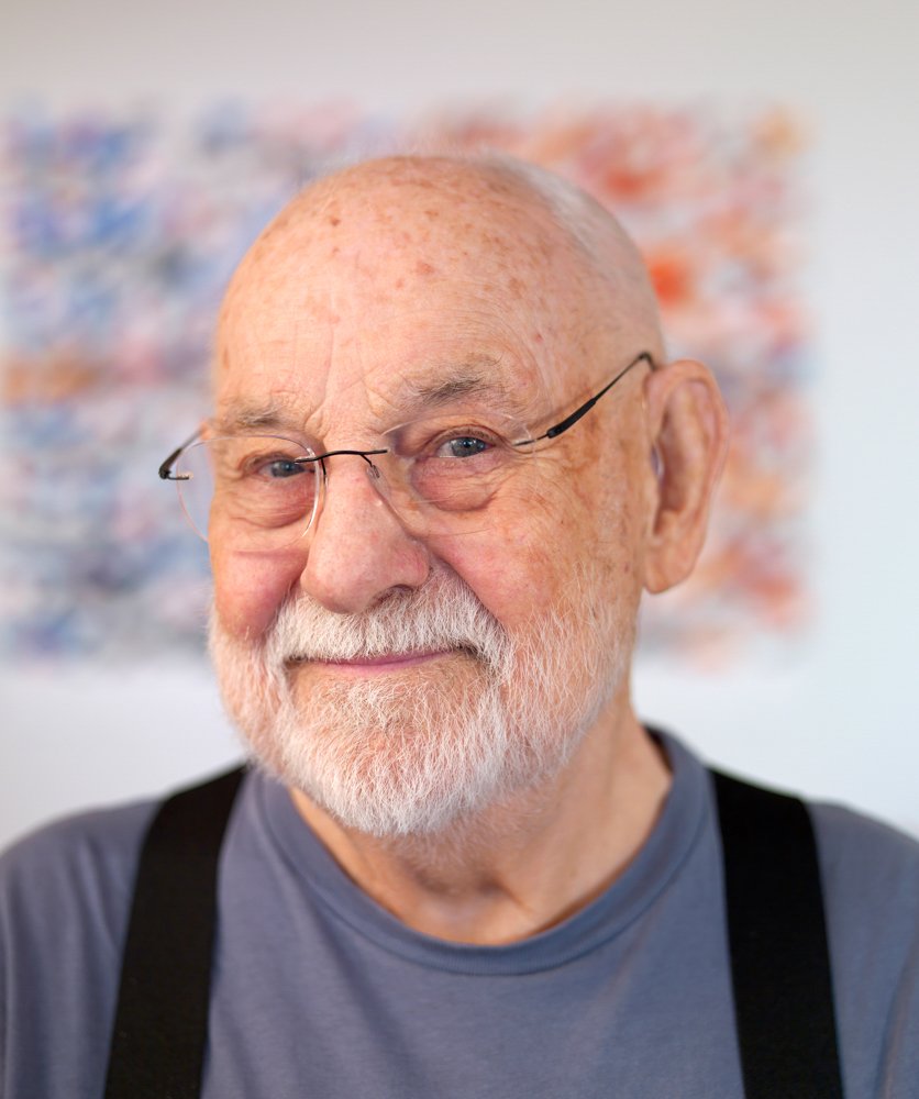 青い瞳でめがねをかけた、80代くらいの男性の、胸までの写真。少し顔を左に傾け、こちらをニコニコとして見ている。髪もひげも白く、ひげは顔の下半分全体をおおっているが、さわやかに整えられている。濃い灰色のTシャツに黒いサスペンダーをつけている。